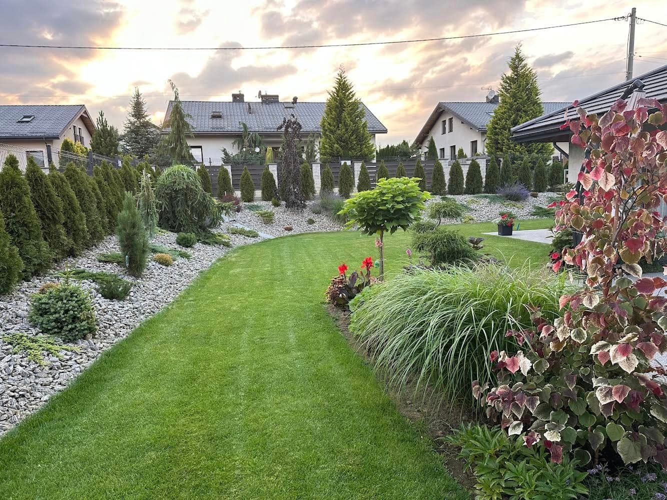 Widok na zadbany ogród z równo przystrzyżonym trawnikiem, ozdobnymi kamieniami, tujami i różnorodną roślinnością, w tle domy jednorodzinne pod pochmurnym niebem.