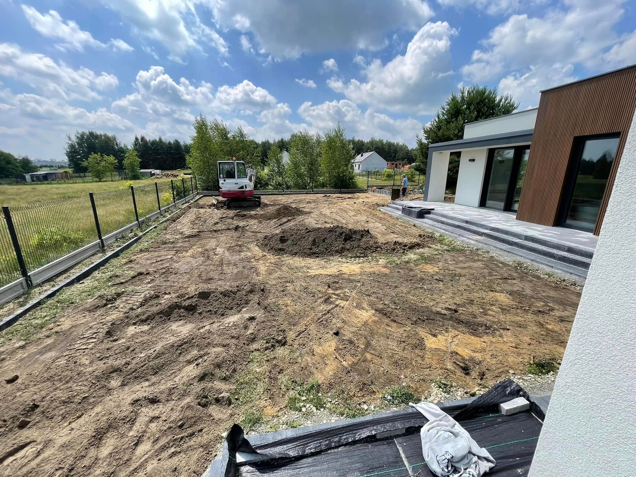 Przygotowanie terenu pod ogród z użyciem mini-koparki, widoczne świeżo wyrównane podłoże, nowoczesny dom w tle, częściowo ułożone obrzeża i siatka ogrodzeniowa.