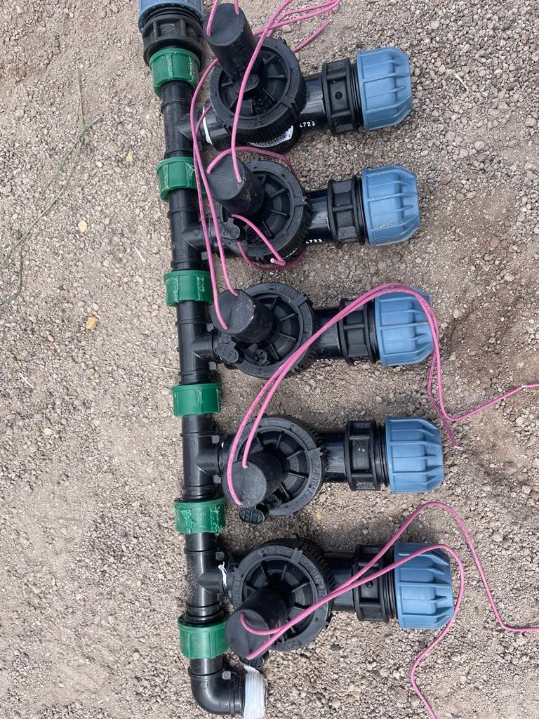 Bliski widok na czarny kolektor z zielonymi złączkami i niebieskimi końcówkami, z podłączonymi przewodami elektrycznymi, leżący na żwirowej powierzchni.