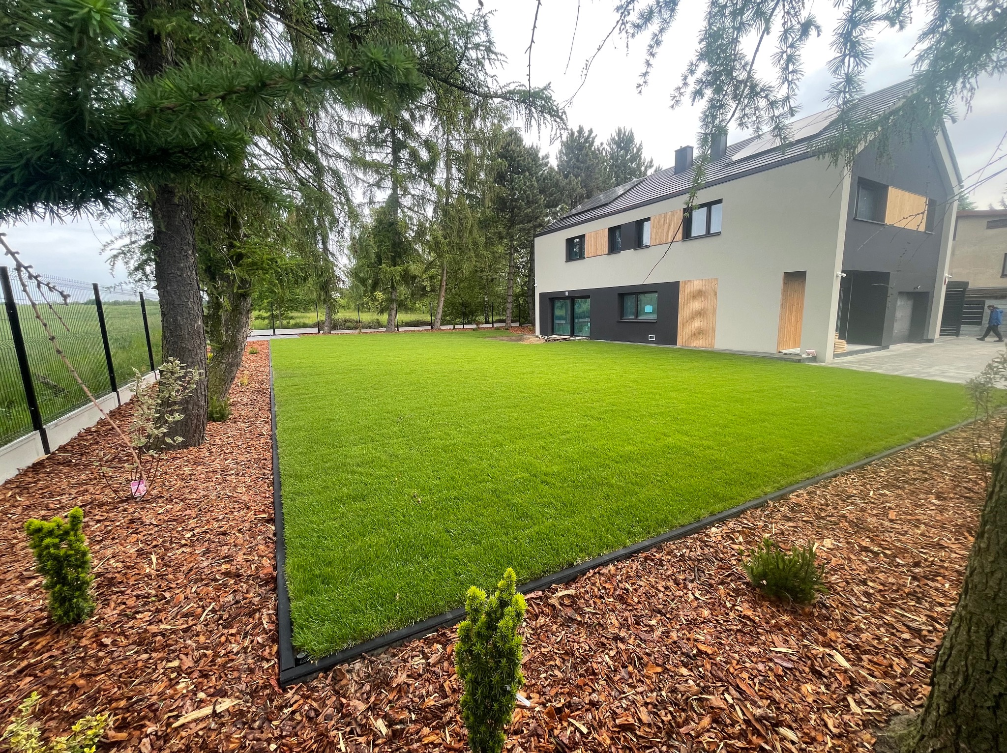 Nowoczesny ogród z intensywnie zielonym trawnikiem, obrzeżem z tworzywa i korą drzewną, na tle domu z elementami drewnianymi i zielenią drzew.