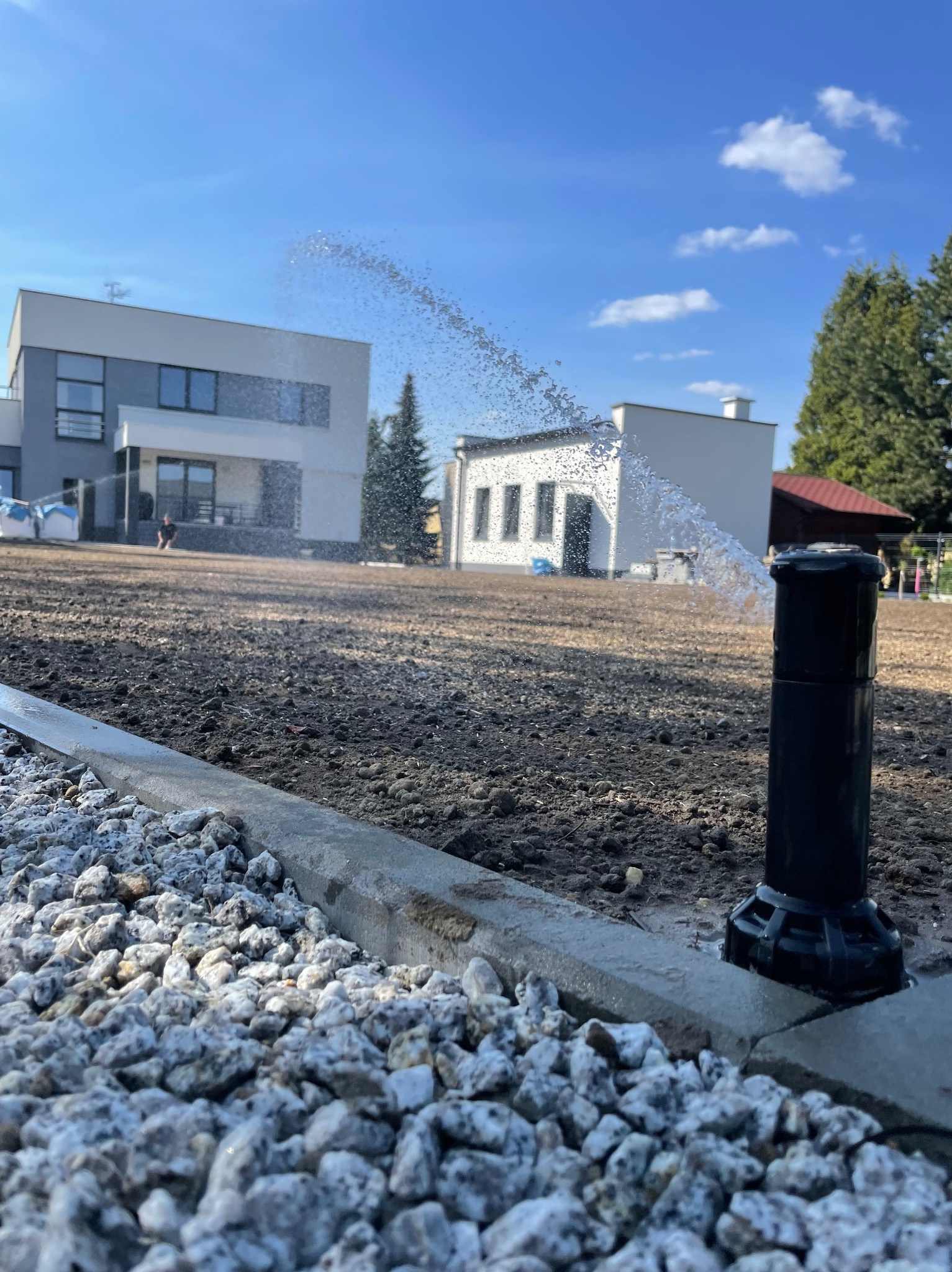 Automatyczny system nawadniania w akcji: zraszacz rozpyla wodę na przygotowany grunt, obok widoczny kamienny obrzeże trawnika i budynki w tle, błękitne niebo.