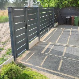 OgrodzeniePanelowe.pl Mateusz Ruszewski - Nowoczesna, szara brama wjazdowa z poziomymi szczeblami i przyległe ogrodzenie panelowe, widoczne ślady spawania na słupkach, na posesji dwa kosze na śmieci.