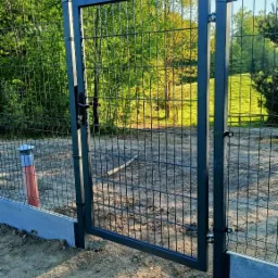 Nowoczesna, czarna brama wjazdowa z metalowej siatki panelowej, zamontowana na betonowej podmurówce, z widocznym lasem w tle.