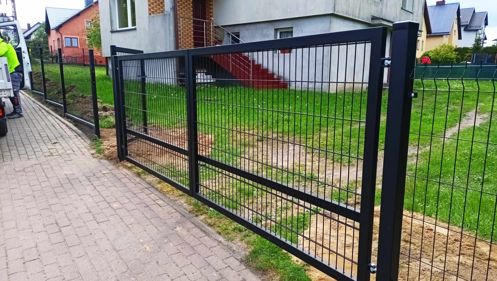 Czarna, metalowa brama wjazdowa z wypełnieniem z paneli ogrodzeniowych, widok z zewnątrz posesji, na pierwszym planie kostka brukowa, w tle trawnik i budynki mieszkalne.