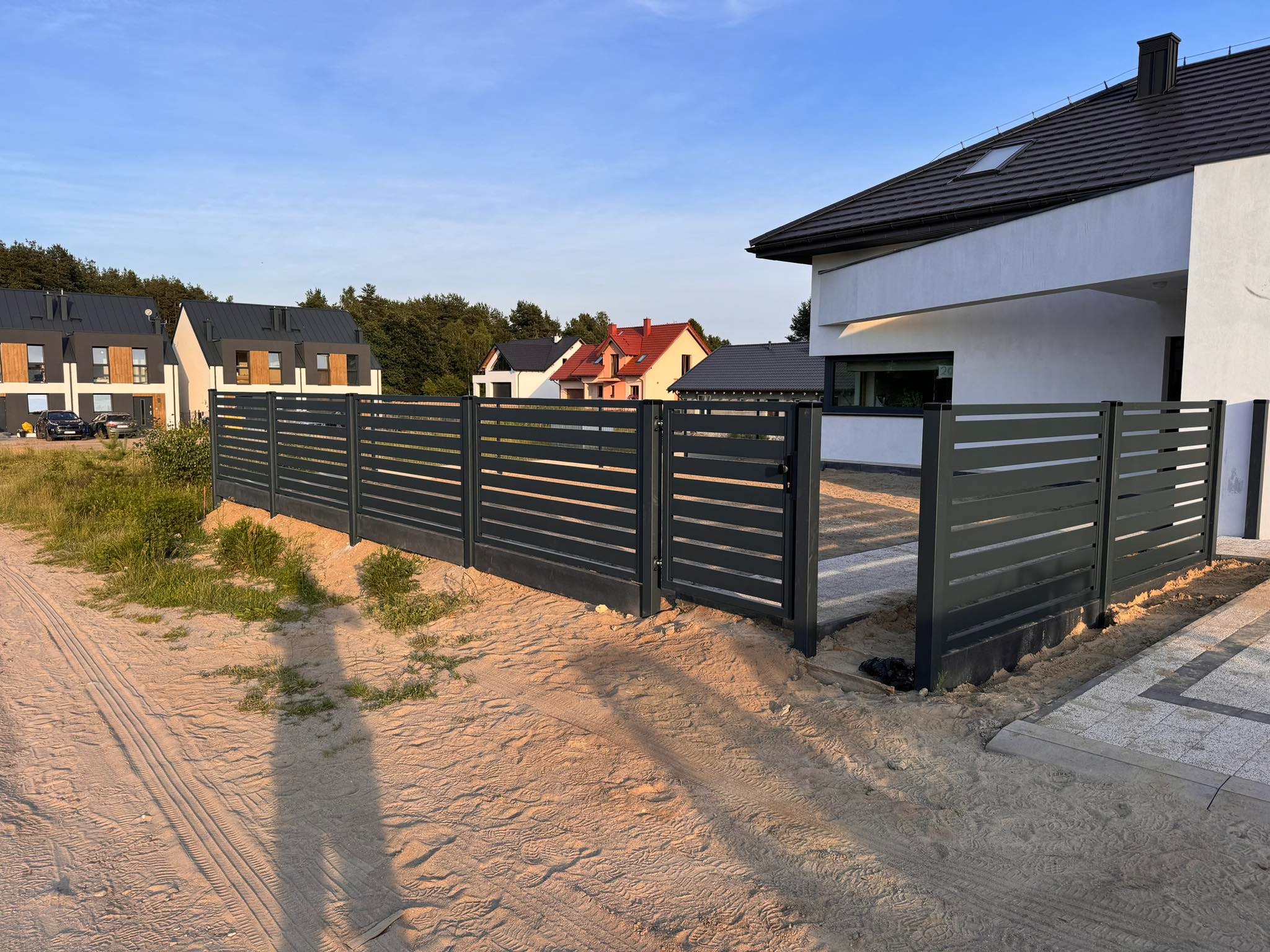 Nowoczesna, pozioma brama wjazdowa w Sierakowicach, z szarymi przęsłami, widok z zewnątrz posesji, na tle jasnego domu z ciemnym dachem i sąsiednich zabudowań.