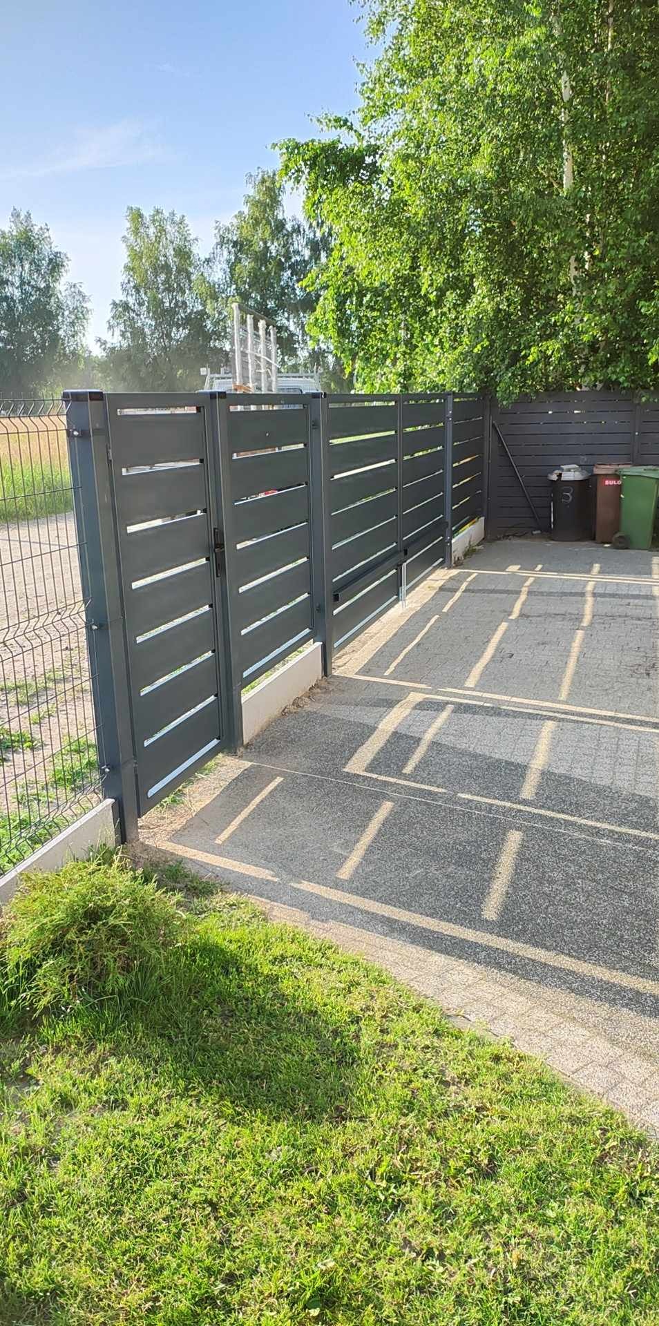 Nowoczesna, szara brama wjazdowa z poziomymi szczeblami i przyległe ogrodzenie panelowe, widoczne ślady spawania na słupkach, na posesji dwa kosze na śmieci.
