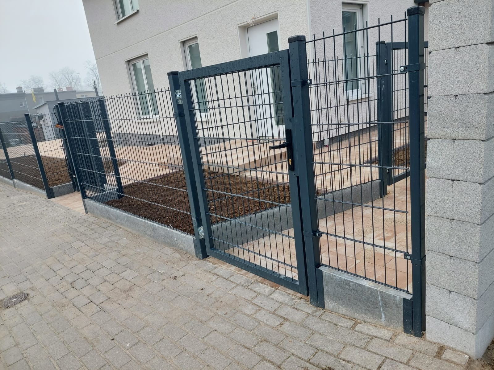 Nowoczesna, grafitowa brama wjazdowa z panelem siatkowym, zamykana na czarny zamek, osadzona na betonowej podmurówce, widok z zewnątrz na posesję z świeżo nawiezioną ziemią.