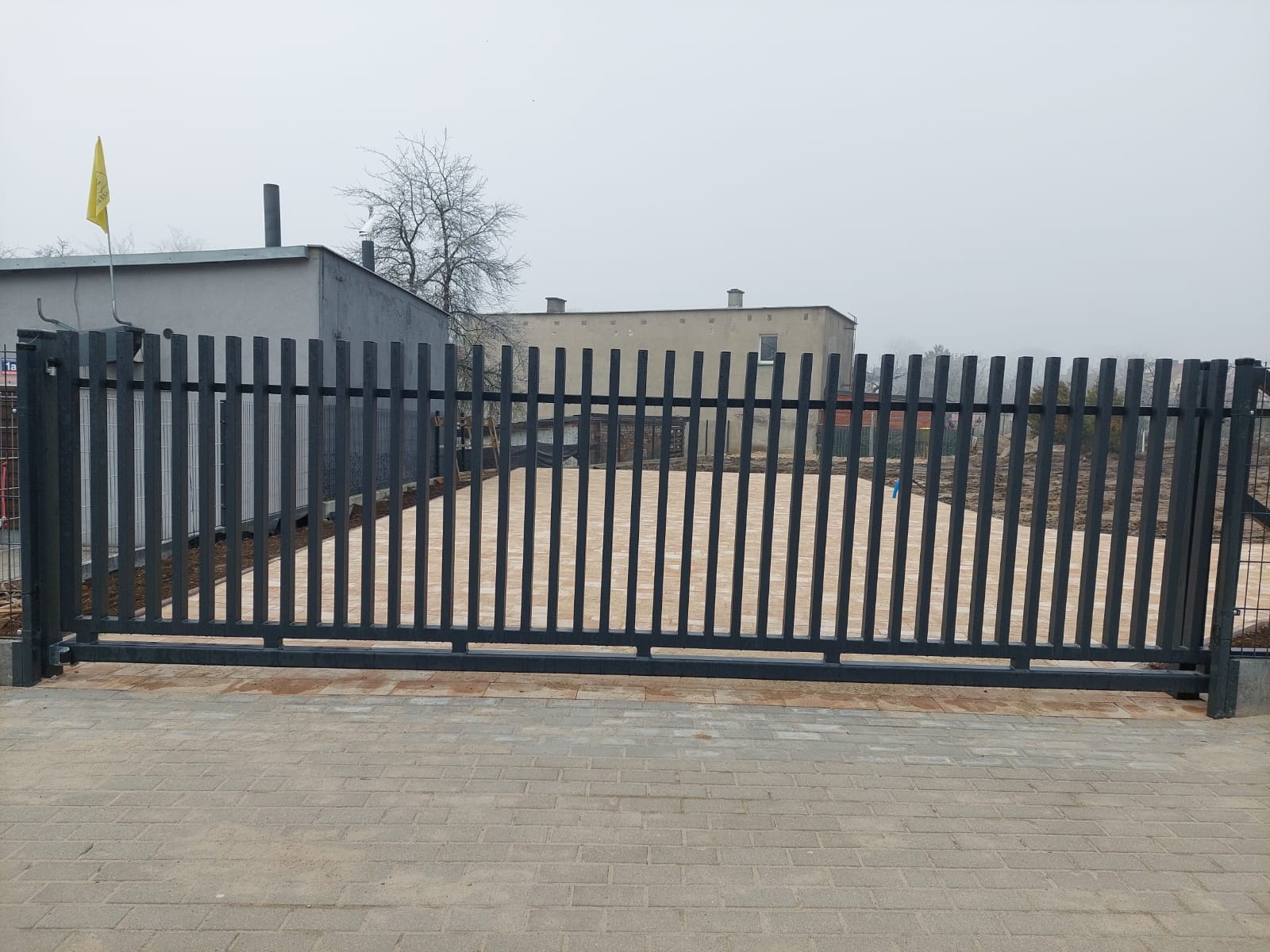Przesuwna brama wjazdowa z pionowych, metalowych profili, widok z zewnątrz na podjazd z kostki brukowej i budynki w tle, pochmurny dzień.