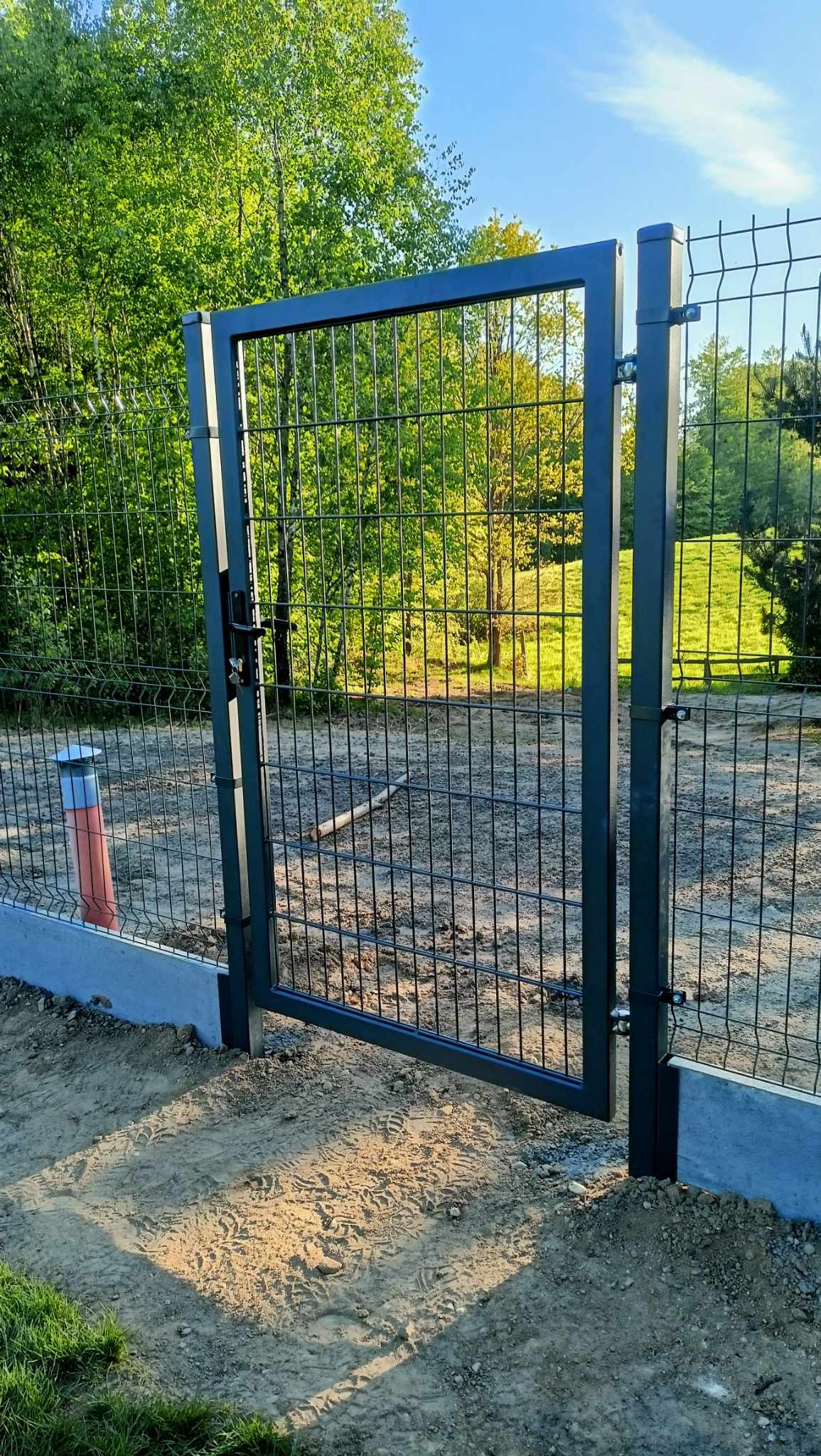 Nowoczesna, czarna brama wjazdowa z metalowej siatki panelowej, zamontowana na betonowej podmurówce, z widocznym lasem w tle.