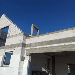 Wznoszenie ścian domu z betonu komórkowego Ytong, widoczne okno wstawione w mur, zbrojenie wieńca i fragment szalunku na tle błękitnego nieba.