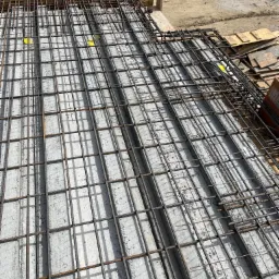 Zbrojenie płyty fundamentowej z betonu, widok z góry, z deskowaniem i materiałami budowlanymi w tle.