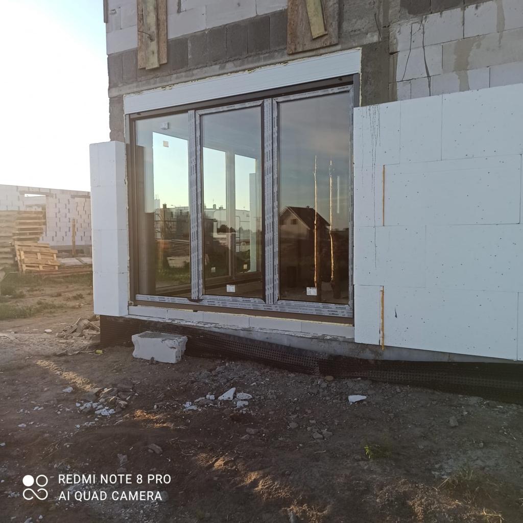 Wznoszenie ściany z bloczków, z zamontowanym oknem tarasowym i częściowo przyklejonymi płytami styropianowymi, widoczne elementy konstrukcyjne i podłoże z gruzem.