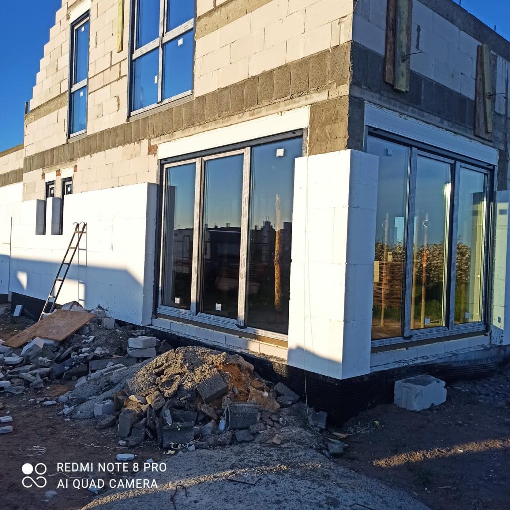Surowy stan domu z betonu komórkowego, z zamontowanymi dużymi oknami przesuwnymi, częściowo ocieplony styropianem, widoczne resztki gruzu i drabina oparta o ścianę.