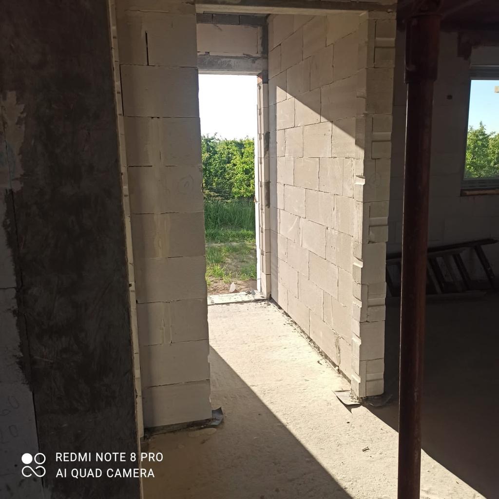 Wnętrze domu w trakcie budowy, widoczne ściany z białych bloczków, surowy beton na podłodze, widok na zewnątrz przez otwór drzwiowy.