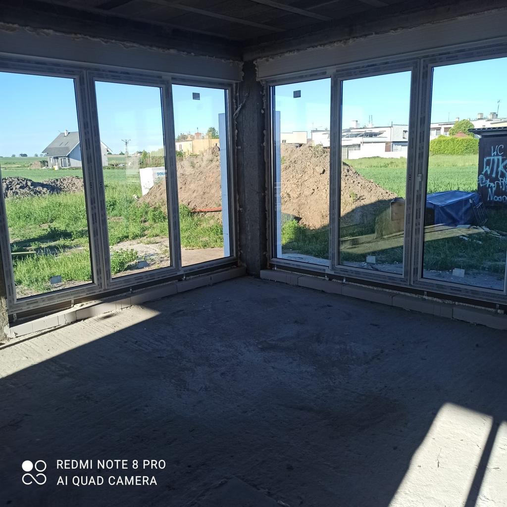 Wnętrze domu w stanie surowym zamkniętym z dużymi oknami PCV, widok na zewnątrz na trawnik i hałdę ziemi, ściany z betonu komórkowego.