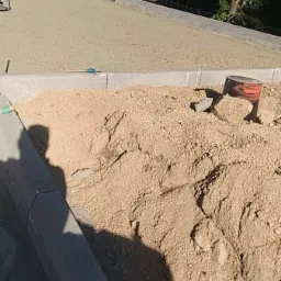 Przygotowanie podłoża pod kostkę brukową w Jeleniej Górze: wyrównany piasek i ustawione obrzeża, widoczny cień osoby oraz ubijak w tle.