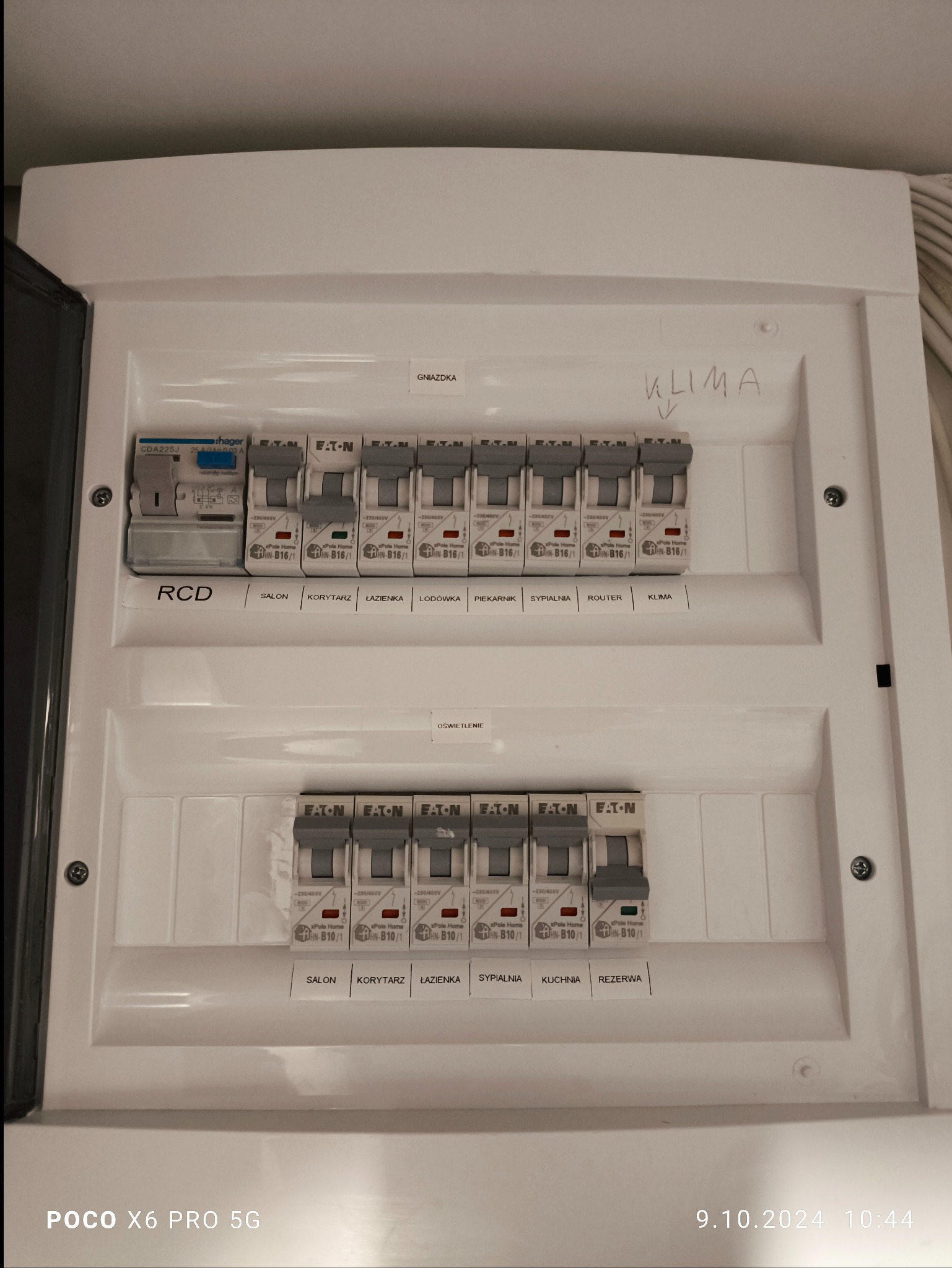 Rozdzielnica elektryczna z bezpiecznikami Eaton, opisana etykietami 'Salon', 'Korytarz', 'Łazienka', 'Lodówka', 'Piekarnik', 'Sypialnia', 'Router', 'Klima', oraz 'Gniazdka' i 'Oświetlenie',...