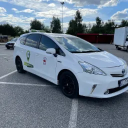 Biała taksówka hybrydowa z czarnymi felgami, zaparkowana na parkingu z widocznym numerem telefonu i logo firmy na drzwiach, na dachu zielony napis TAXI.