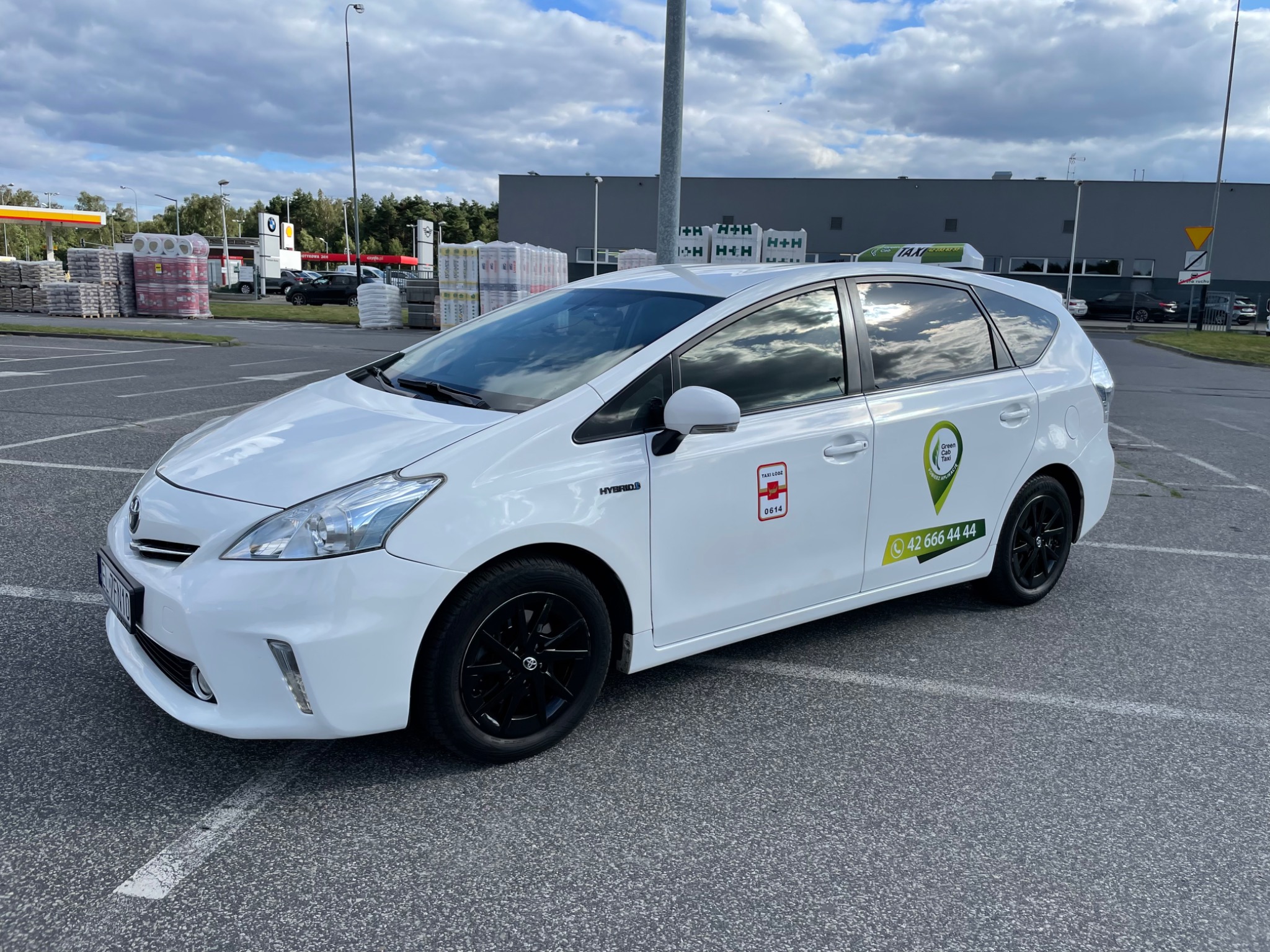Biała Toyota Prius, samochód taksówkarski z czarnymi felgami, zaparkowany na parkingu z widocznymi oznaczeniami Taxi Łódź i numerem telefonu, na tle sklepu budowlanego i pochmurnego nieba.