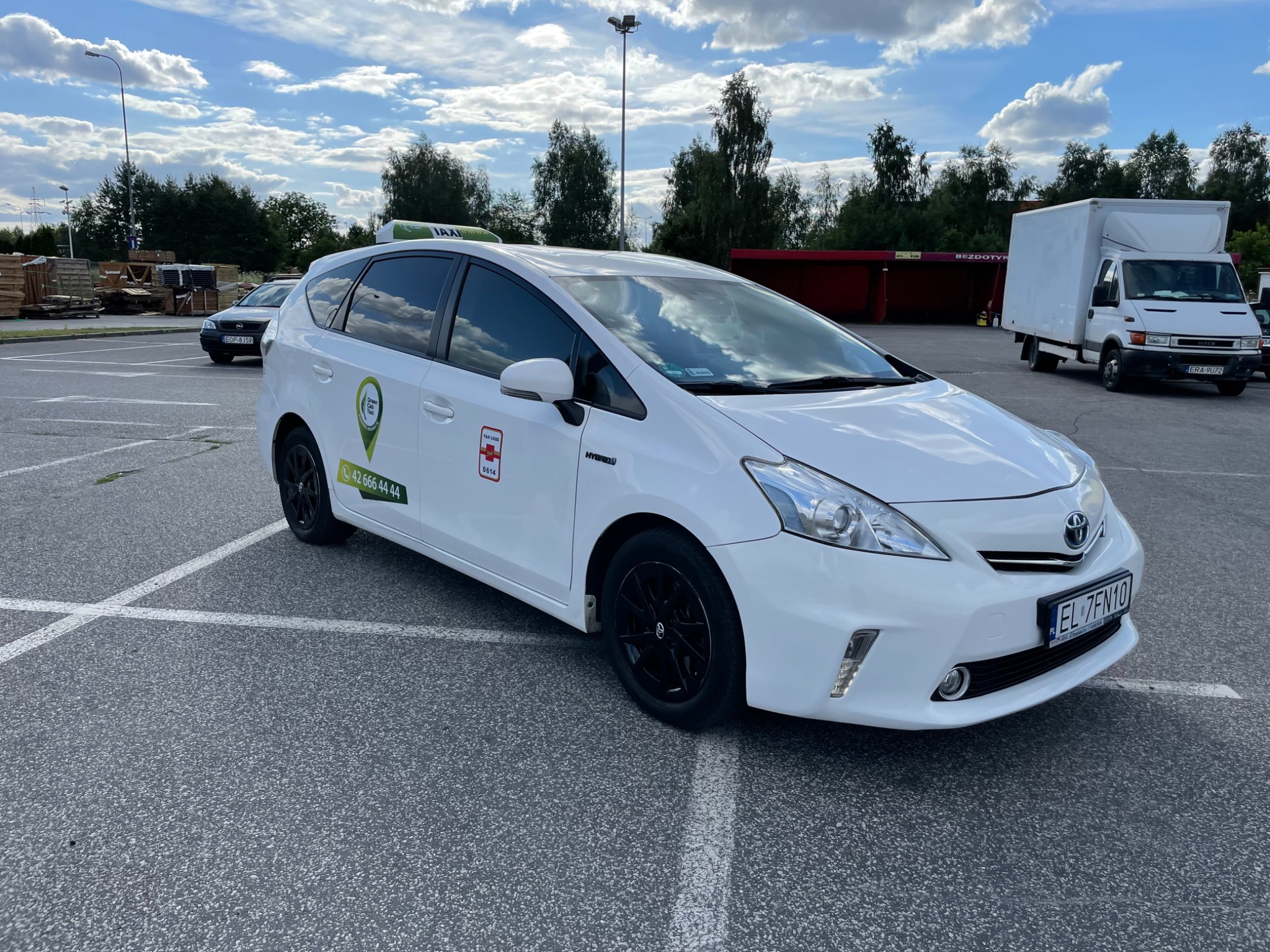 Biała taksówka hybrydowa z czarnymi felgami, zaparkowana na parkingu z widocznym numerem telefonu i logo firmy na drzwiach, na dachu zielony napis TAXI.