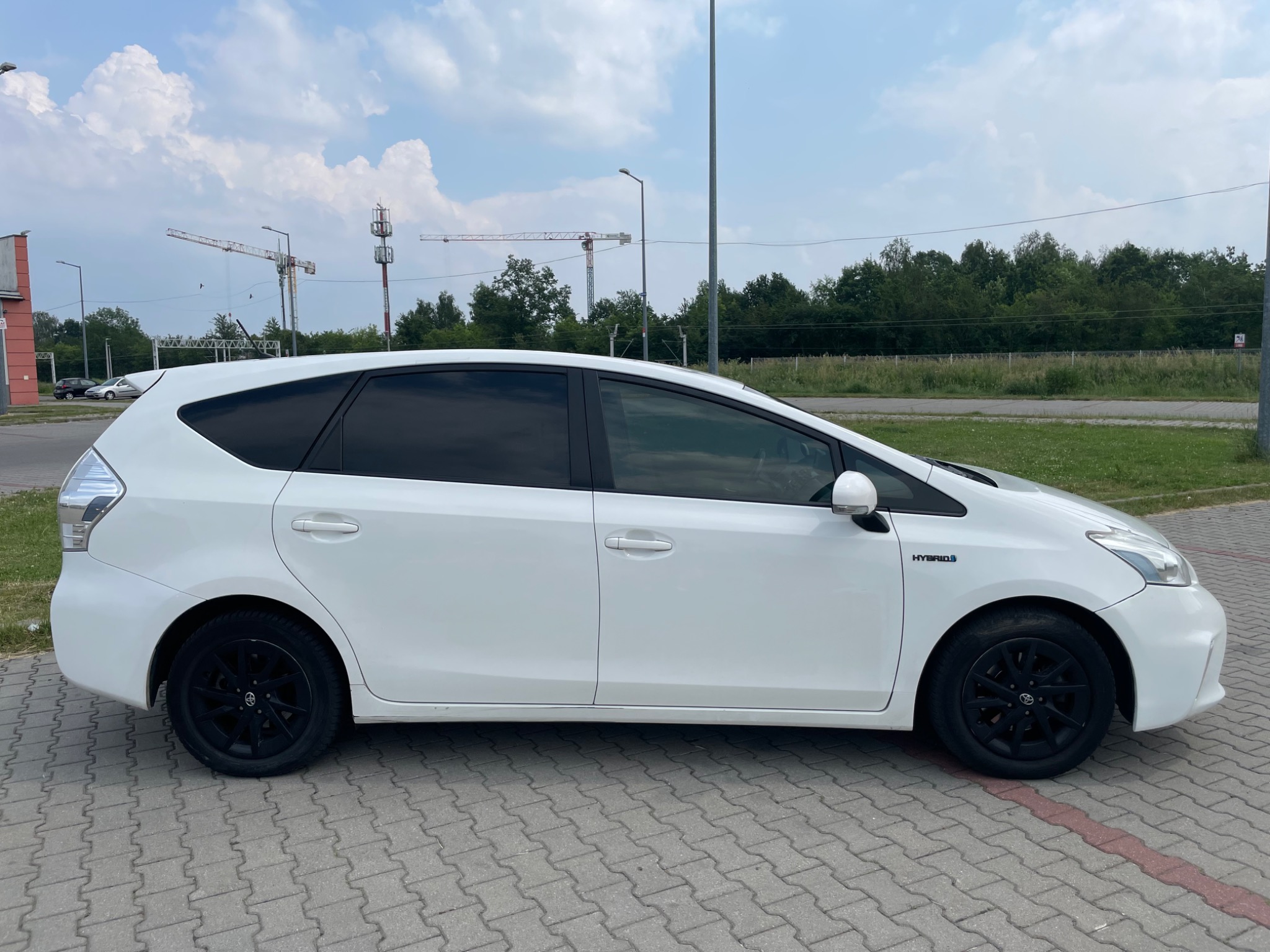 Biały samochód hybrydowy z czarnymi felgami, zaparkowany na kostce brukowej, w tle słupy wysokiego napięcia i żurawie budowlane.