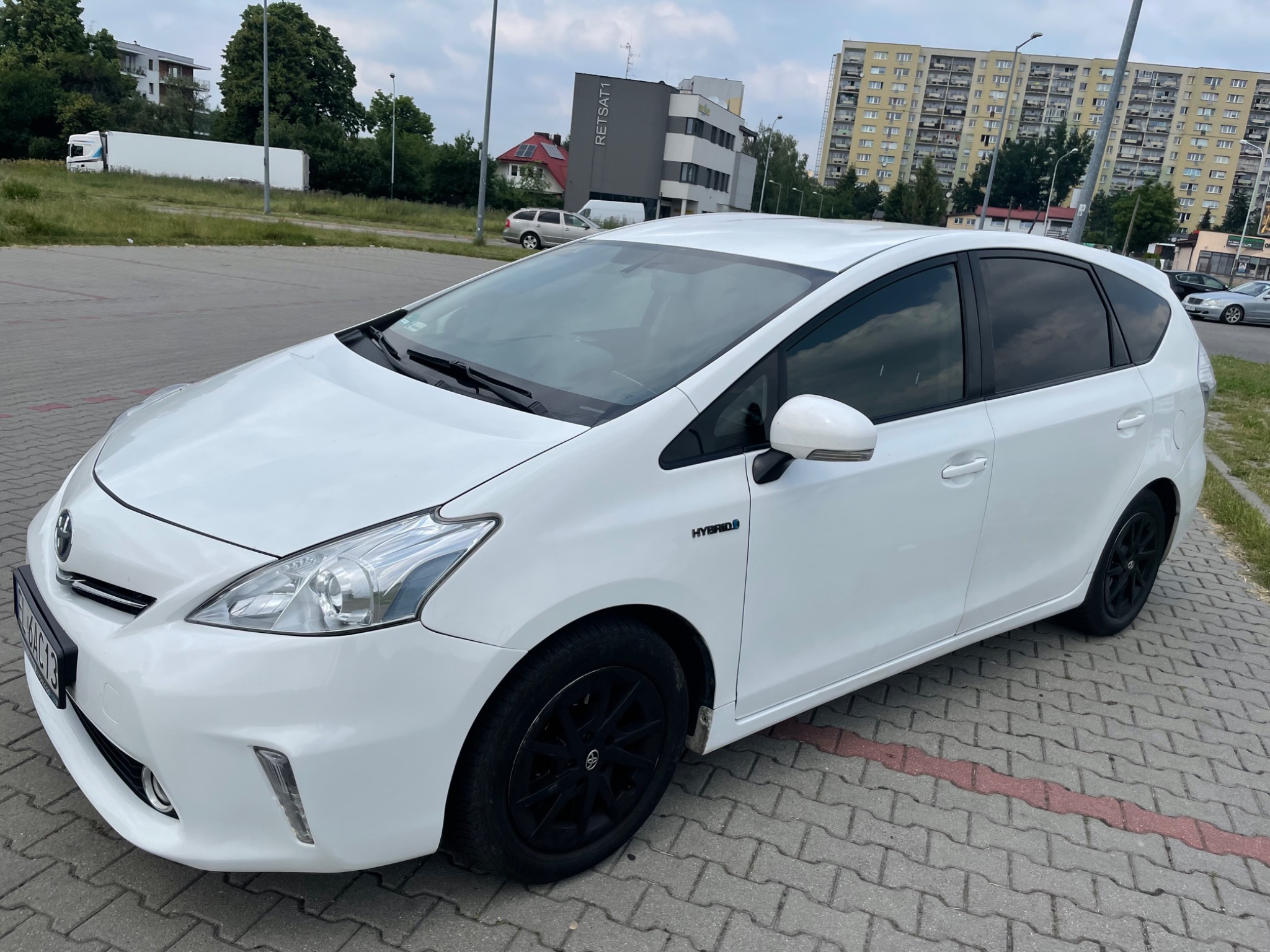 Biała hybryda z czarnymi felgami zaparkowana na szarym bruku, w tle budynki mieszkalne i biurowe.