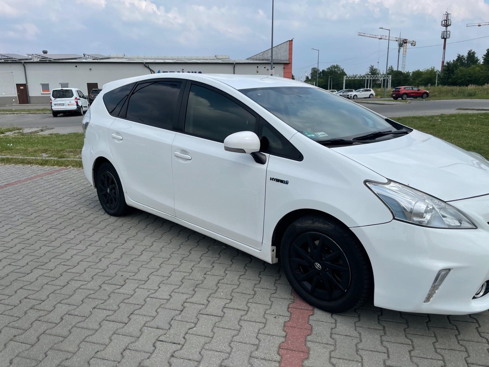 Biały samochód hybrydowy z czarnymi felgami, zaparkowany na kostce brukowej w Łodzi, z widocznym napisem 'Hybrid' na błotniku.