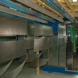 Elementy metalowe zawieszone na linii produkcyjnej przed procesem malowania, widoczna kabina lakiernicza i system transportu detali.