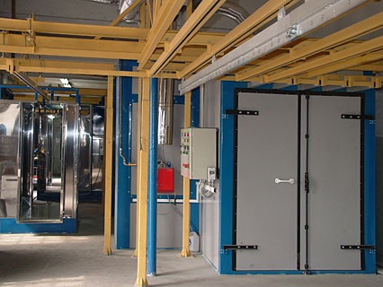 Wnętrze hali z linią technologiczną do obróbki metali, widoczna kabina lakiernicza z dwuskrzydłowymi drzwiami i system transportu podwieszanego z elementami w trakcie procesu.