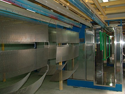 Elementy metalowe zawieszone na linii produkcyjnej przed procesem malowania, widoczna kabina lakiernicza i system transportu detali.