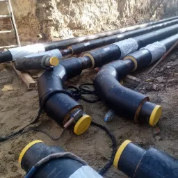 Czarne, izolowane rury z żółtymi zaślepkami ułożone w wykopie ziemnym podczas instalacji, widoczne narzędzia i materiały izolacyjne.