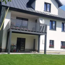 Dwukondygnacyjny dom z balkonem z metalową balustradą, satelitą na ścianie i zadbanym trawnikiem.