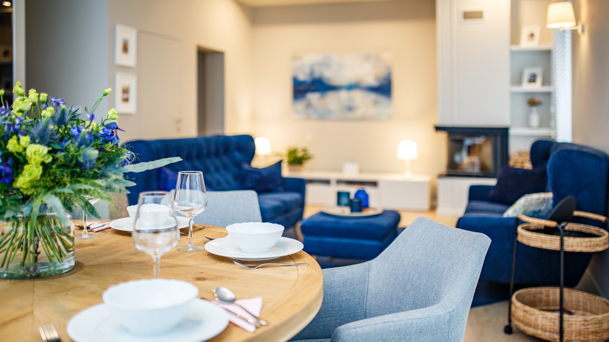 Jasne wnętrze salonu z jadalnią: okrągły drewniany stół z nakryciem, kwiaty w wazonie, w tle niebieska sofa i fotel, kominek.