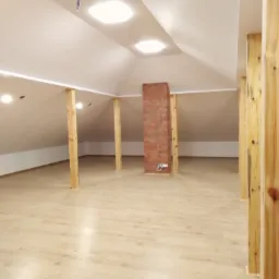 Poddasze po remoncie z drewnianymi słupami, wykończone panelami podłogowymi i ceglanym kominem, oświetlone wpuszczanymi lampami.