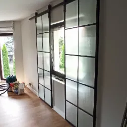 Przesuwne drzwi loftowe z czarnymi szprosami i matowym szkłem, zamontowane na szynie, widoczne z boku w jasnym wnętrzu. W tle okno z widokiem na zieleń i drabina.