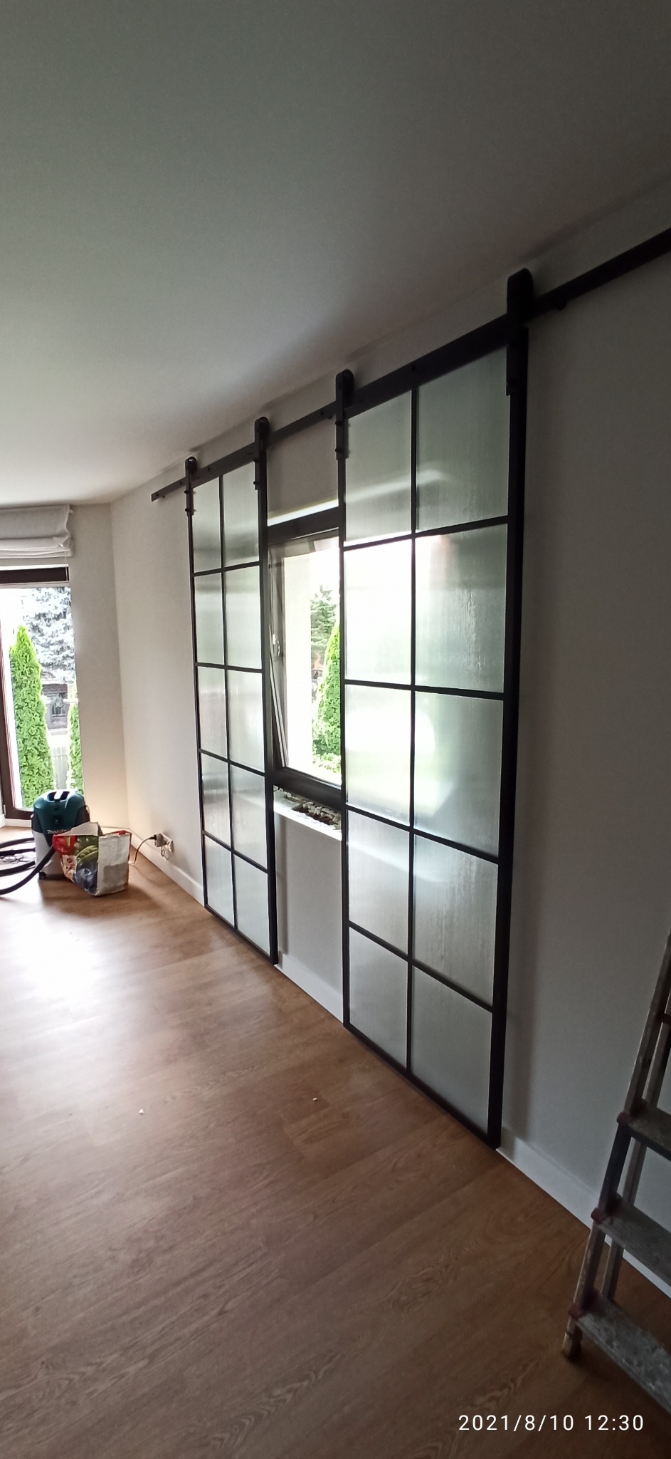 Przesuwne drzwi loftowe z czarnymi szprosami i matowym szkłem, zamontowane na szynie, widoczne z boku w jasnym wnętrzu. W tle okno z widokiem na zieleń i drabina.