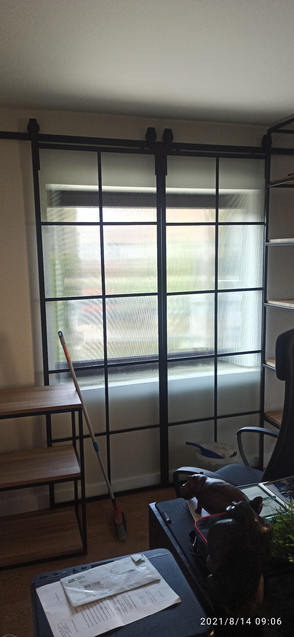 Przesuwne drzwi wewnętrzne z czarną metalową ramą i teksturowanym szkłem, widoczne z wnętrza pomieszczenia, z mopem opartym o ramę i elementami wyposażenia wnętrz w tle.