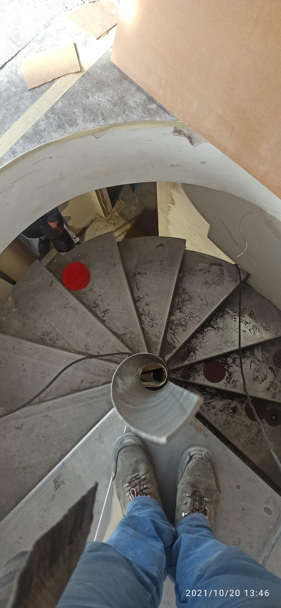 Wnętrze spiralnej klatki schodowej w trakcie montażu, widok z góry na stalowe stopnie, fragment rury w centrum i buty osoby stojącej na schodach.
