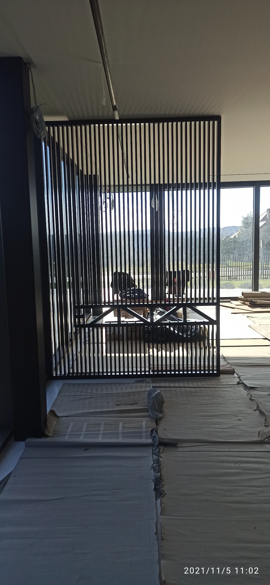 Metalowa konstrukcja z pionowych listew, prawdopodobnie element balustrady lub poręczy, zamontowana w nowoczesnym wnętrzu z dużymi oknami i widokiem na zewnątrz, podłoga zabezpieczona folią malarską.