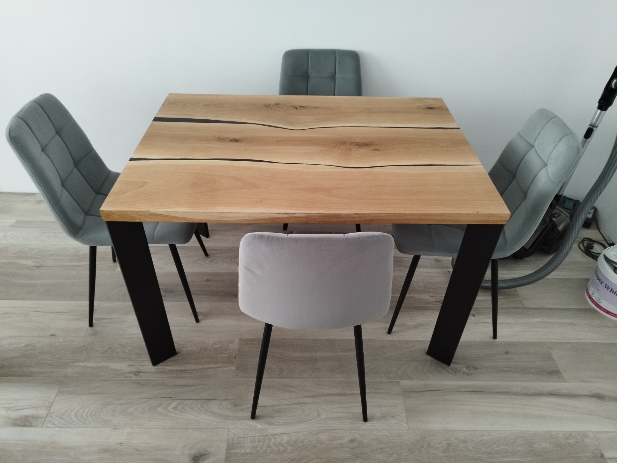 Stół z blatem z jasnego drewna z czarnymi żywicznymi wypełnieniami, wsparty na czarnych, prostokątnych nogach, otoczony czterema szarymi krzesłami z tapicerowanymi siedziskami i oparciami.