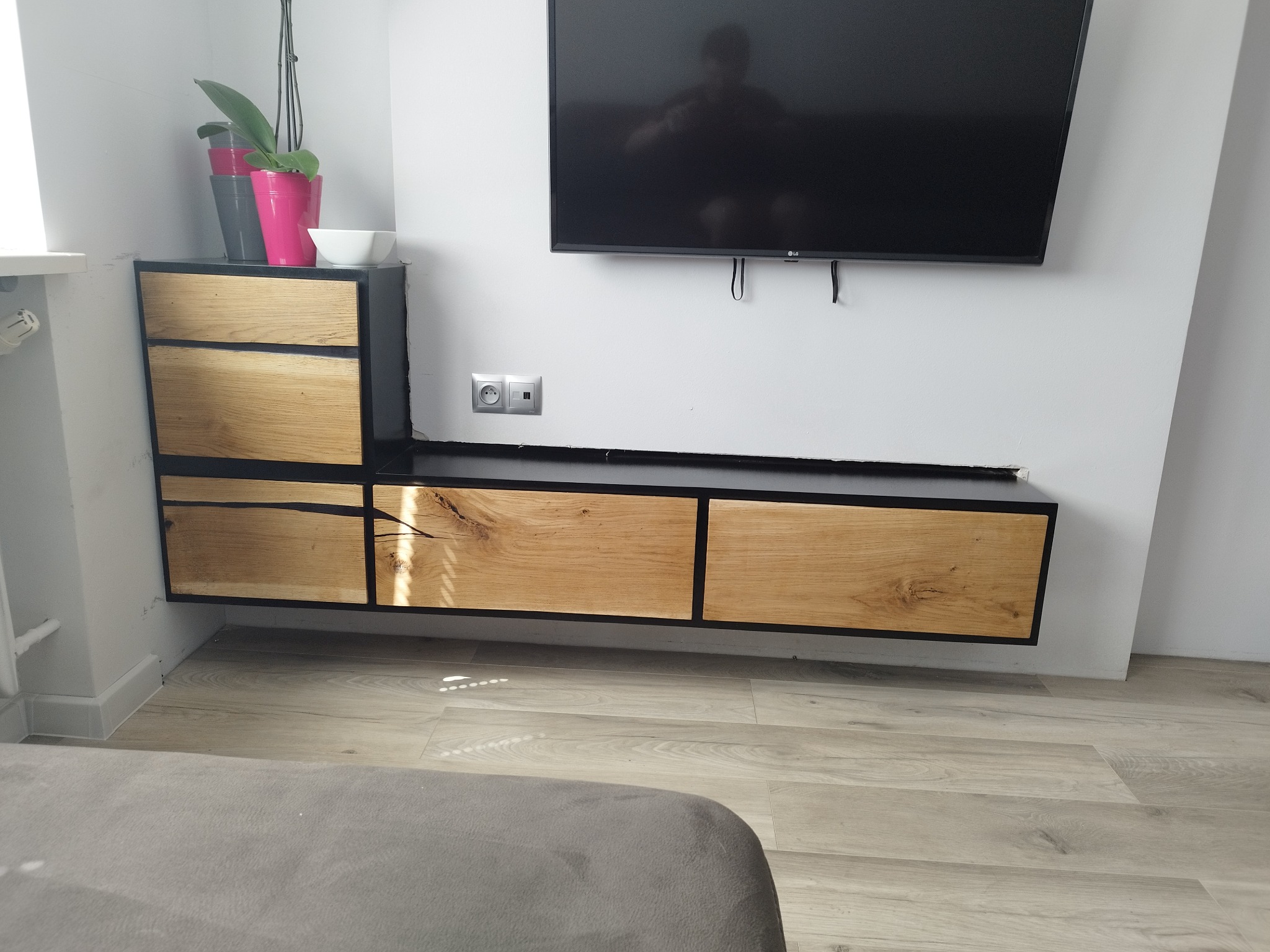 Nowoczesna szafka RTV z drewna i metalu, zawieszona na ścianie pod telewizorem, z widocznymi elementami dekoracyjnymi: doniczki z kwiatami i miseczka.