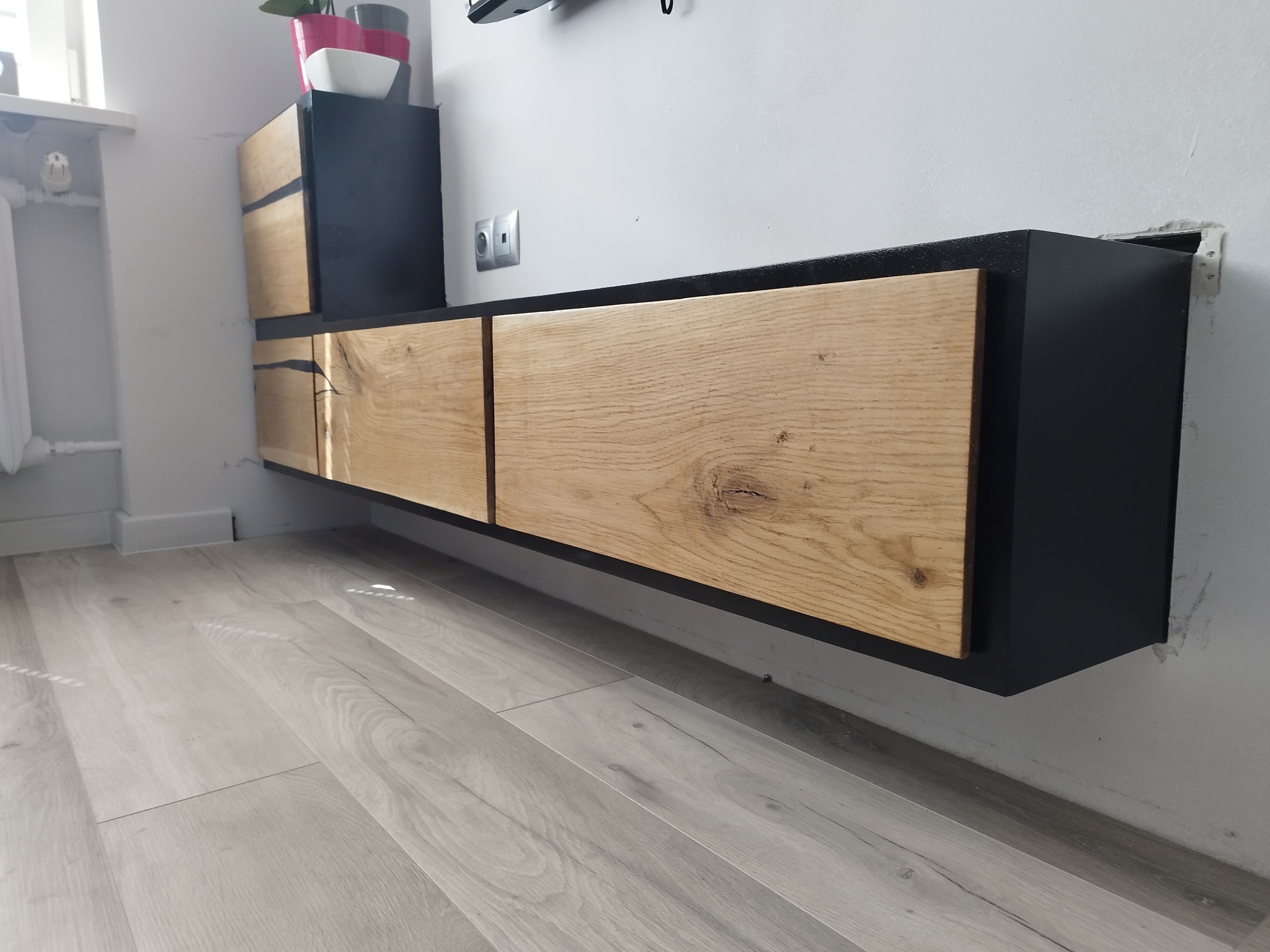 Nowoczesna szafka RTV wisząca, z frontami z jasnego drewna o wyraźnym usłojeniu i czarną obudową, zamontowana na szarej ścianie nad panelami podłogowymi w odcieniach szarości.