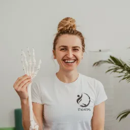 Uśmiechnięta kobieta w białej koszulce z logo Pilates trzyma w dłoni model szkieletu dłoni, w tle roślina doniczkowa i białe ściany.