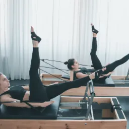 Dwie kobiety w czarnych strojach ćwiczą na drewnianych reformerach pilates, unosząc nogi w powietrze w jasnym studio z zasłonami.