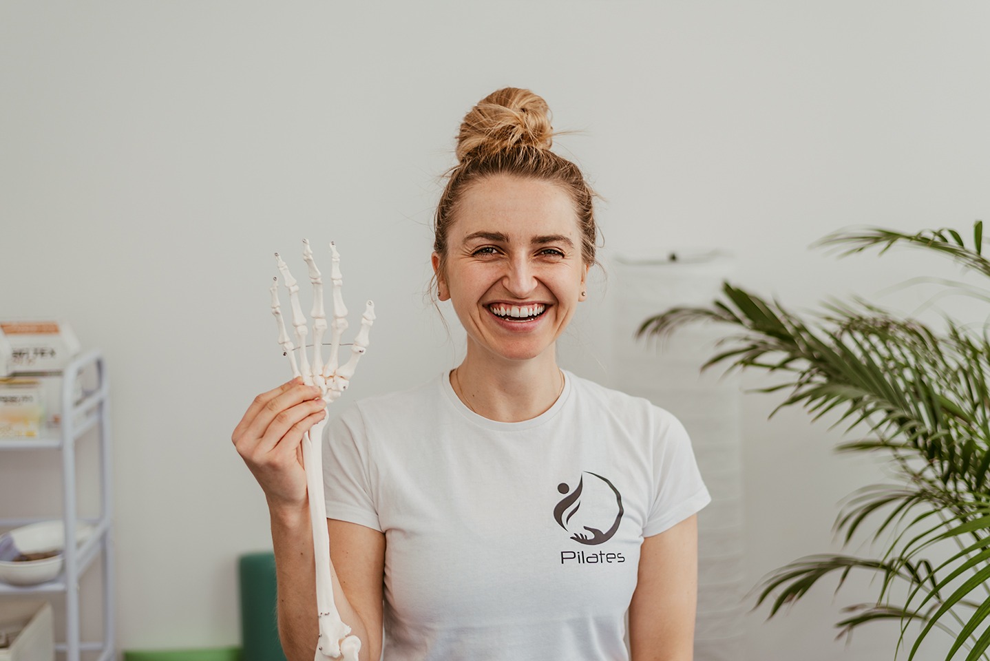 Uśmiechnięta kobieta w białej koszulce z logo Pilates trzyma w dłoni model szkieletu dłoni, w tle roślina doniczkowa i białe ściany.