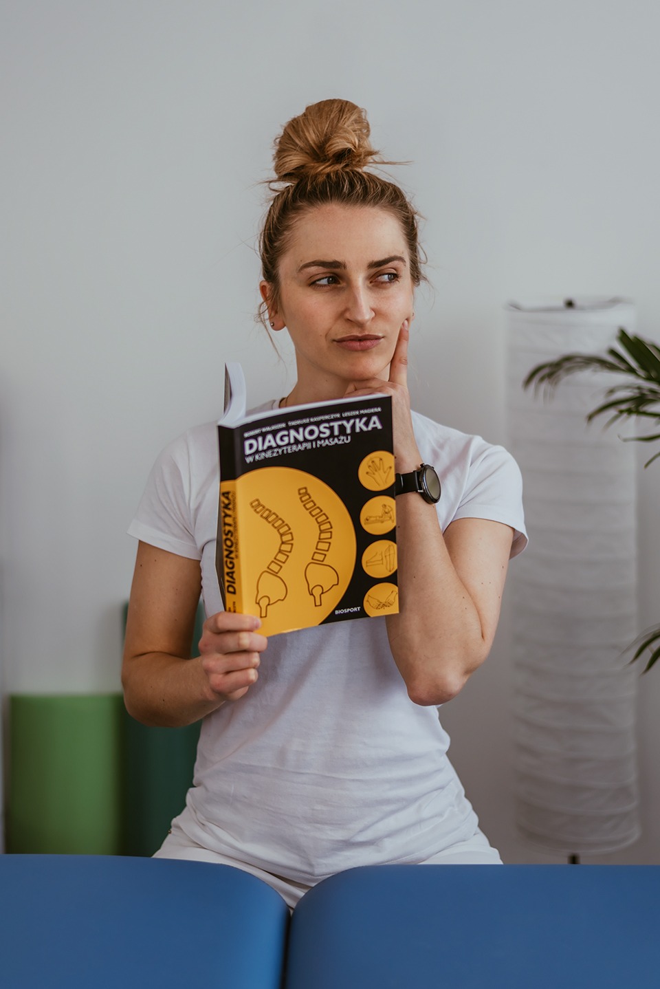 Kobieta z upiętymi włosami, w białym T-shircie, trzyma książkę o diagnostyce w kinezyterapii i masażu, zamyślona, z ręką przy twarzy, na tle gabinetu z elementami wyposażenia.