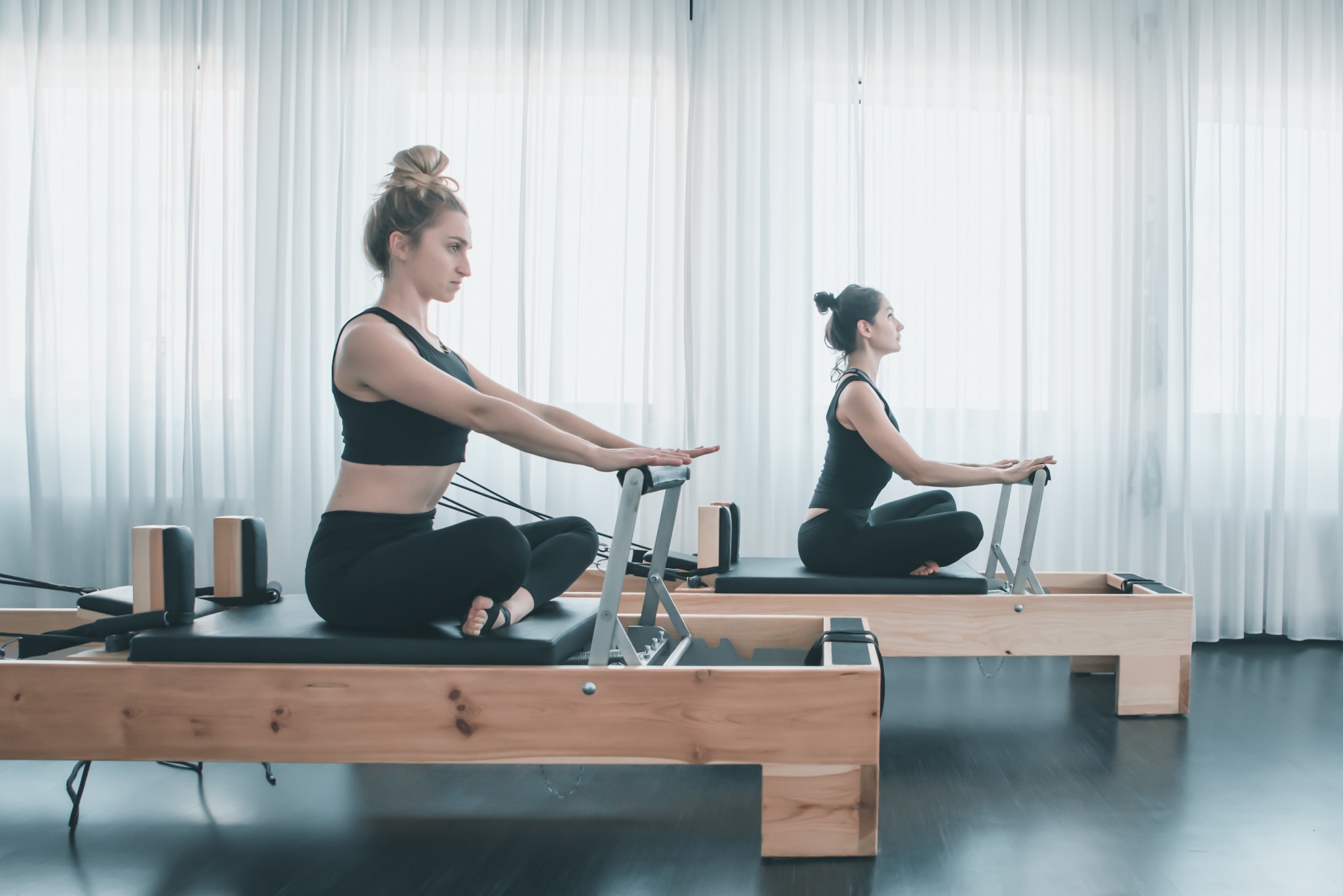 Dwie kobiety ćwiczą na reformerach do pilates w jasnym studio z białymi zasłonami w tle.
