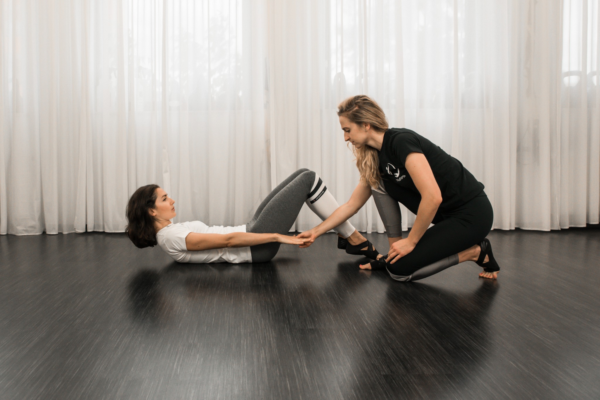 Instruktorka pomaga kobiecie w ćwiczeniach rozciągających na podłodze w studiu we Włocławku, widoczne białe zasłony w tle.