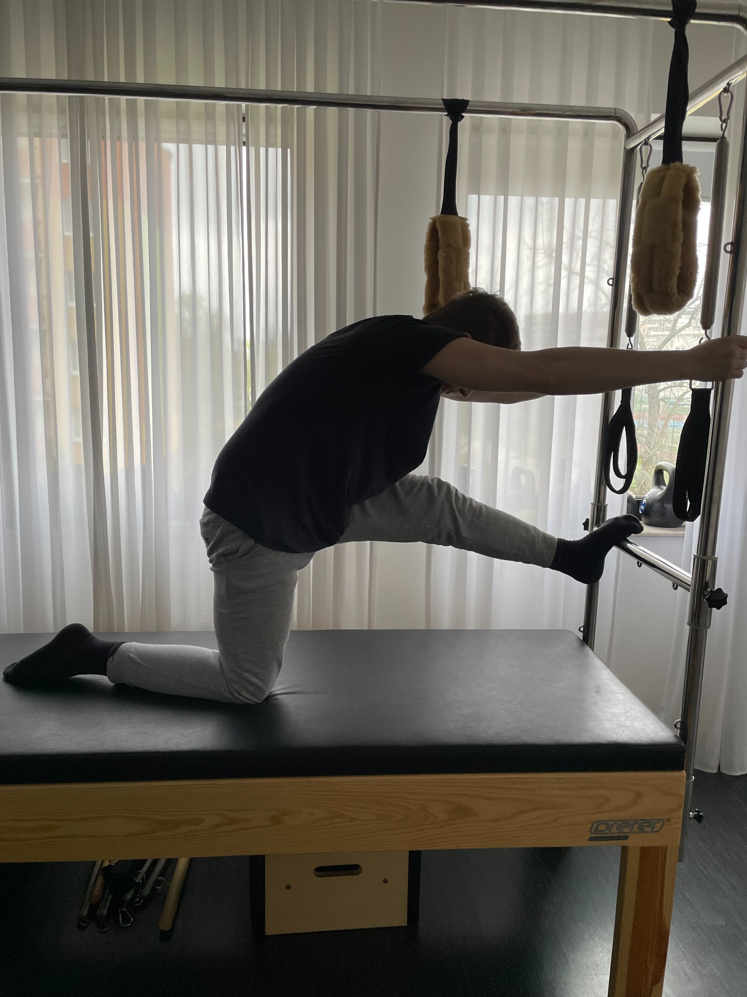 Osoba w czarnej koszulce i szarych spodniach wykonuje ćwiczenie rozciągające na reformerze pilates w jasnym pomieszczeniu z białymi zasłonami.