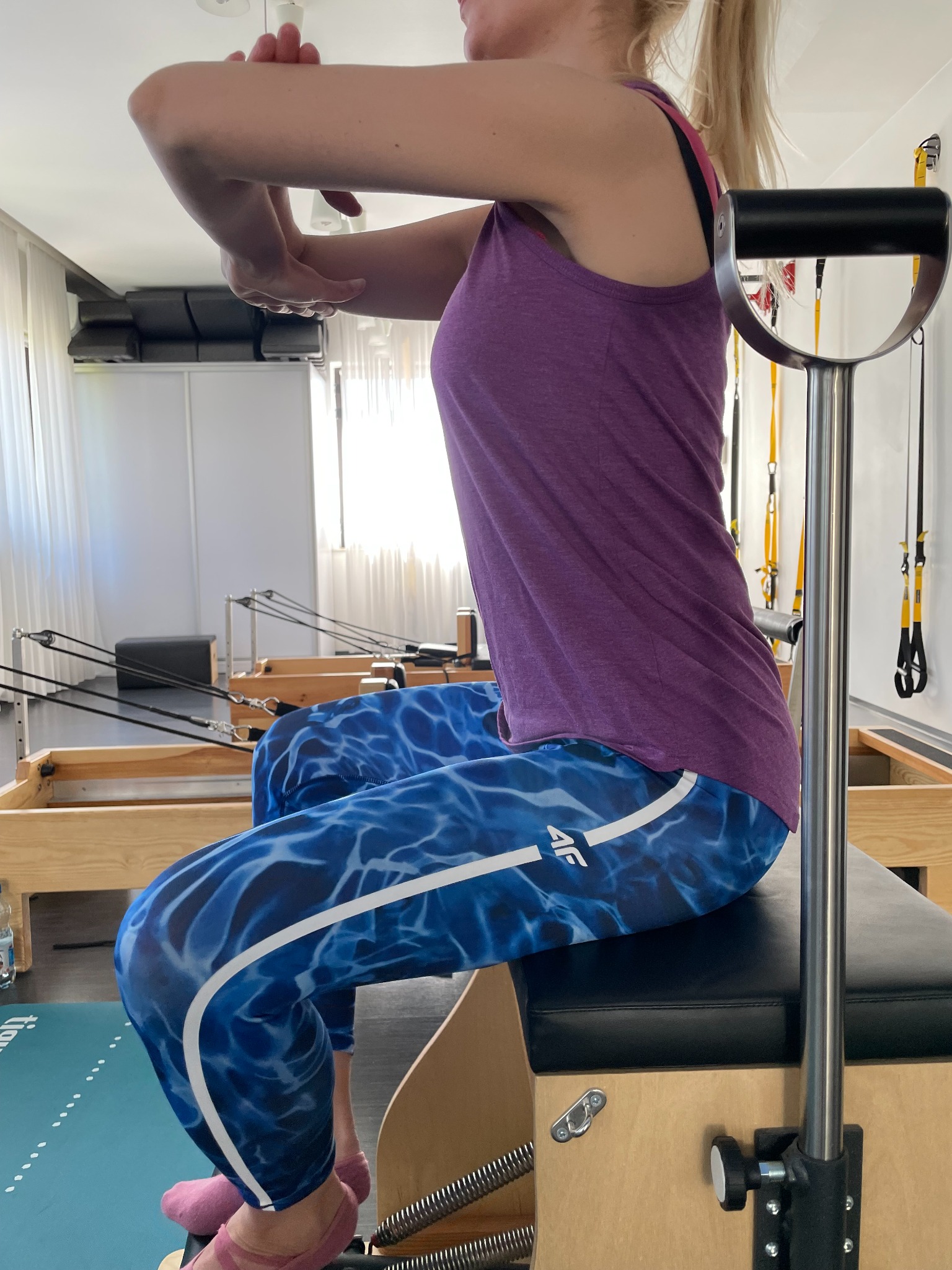 Kobieta w sportowych legginsach i fioletowym topie ćwiczy na reformerze do pilatesu w jasnym studiu.